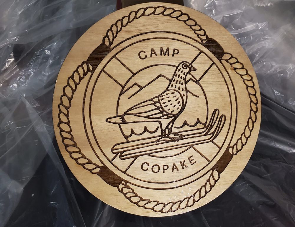 Round wooden plaque engraved with a camp logo, bird and rope border